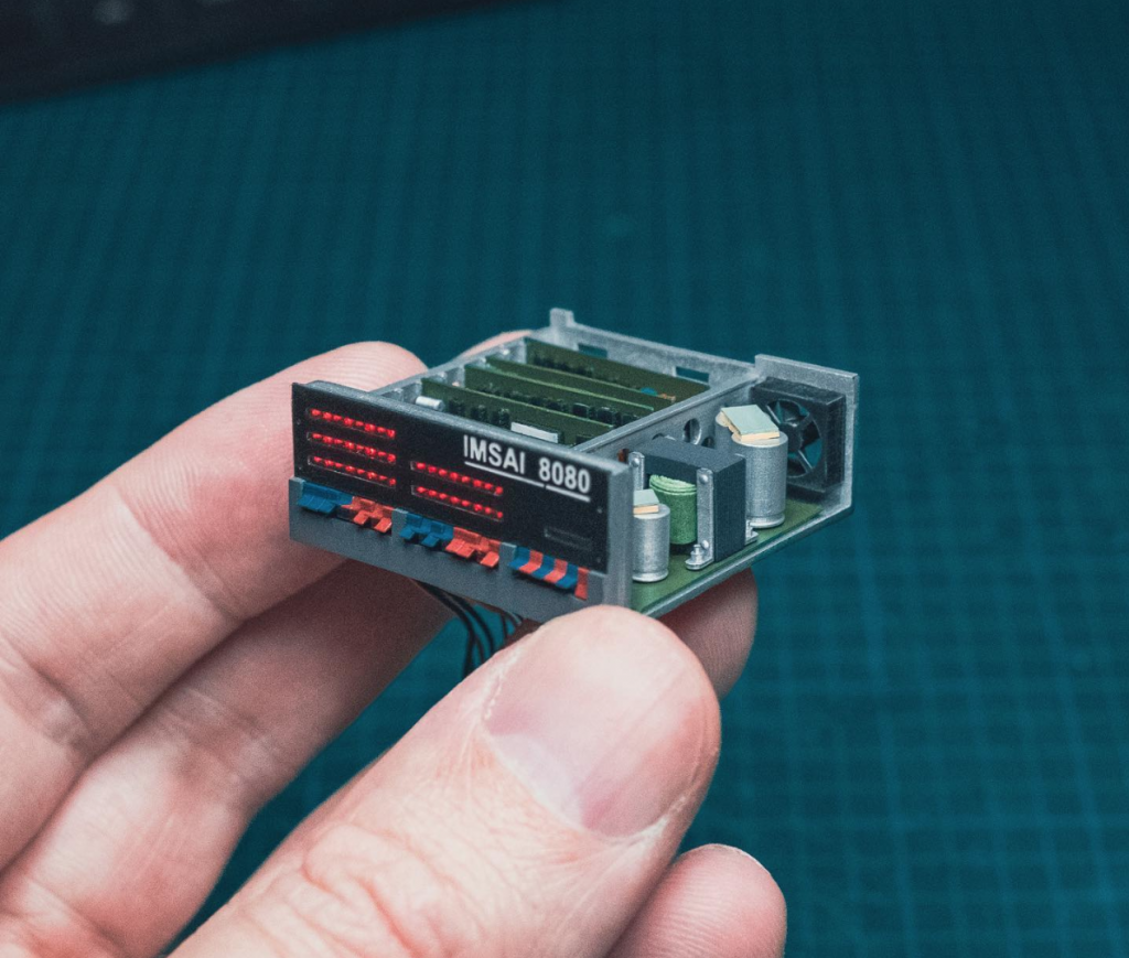 Macro photo of artist's hand holding miniature IMSAI microcomputer, open with exposed boards, fan, and chips