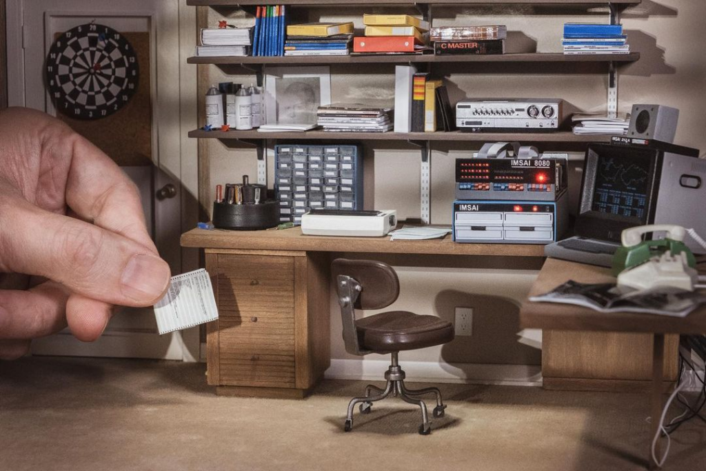 Detailed diorama of David Lightman's bedroom from WarGames with desk, chairs, shelves, and various miniature props, with a hand reaching into frame holding a printout