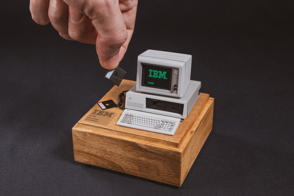 Miniature IBM 5150 PC computer, keyboard, and monitor reading "IBM" on a wooden display stand, with artist's hand reaching into frame holding a tiny floppy disk